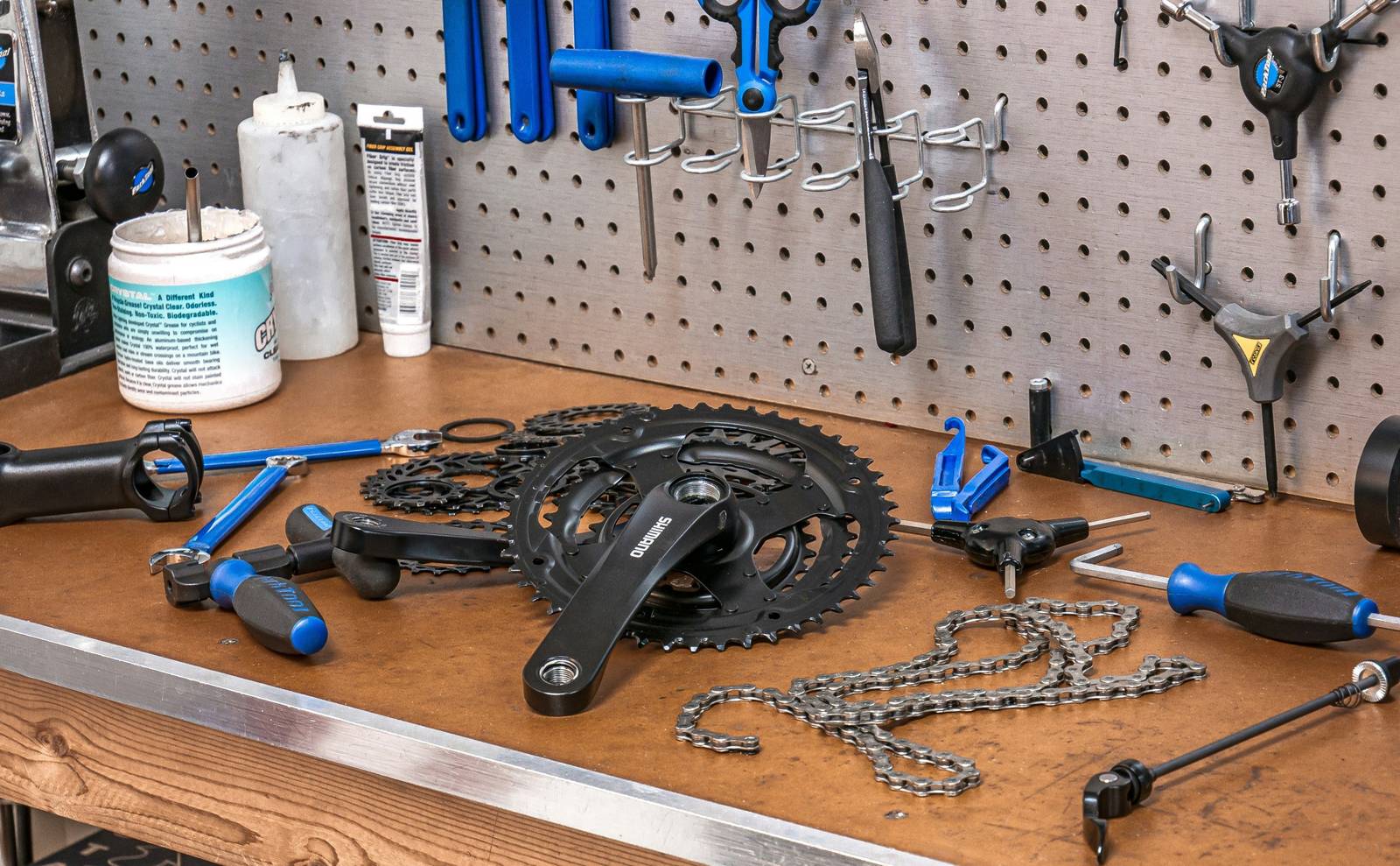 A bike shop workbench with wrenches and hex keys hanging on a pegboard and a crankset, cogs and a chain laid out on the bench.